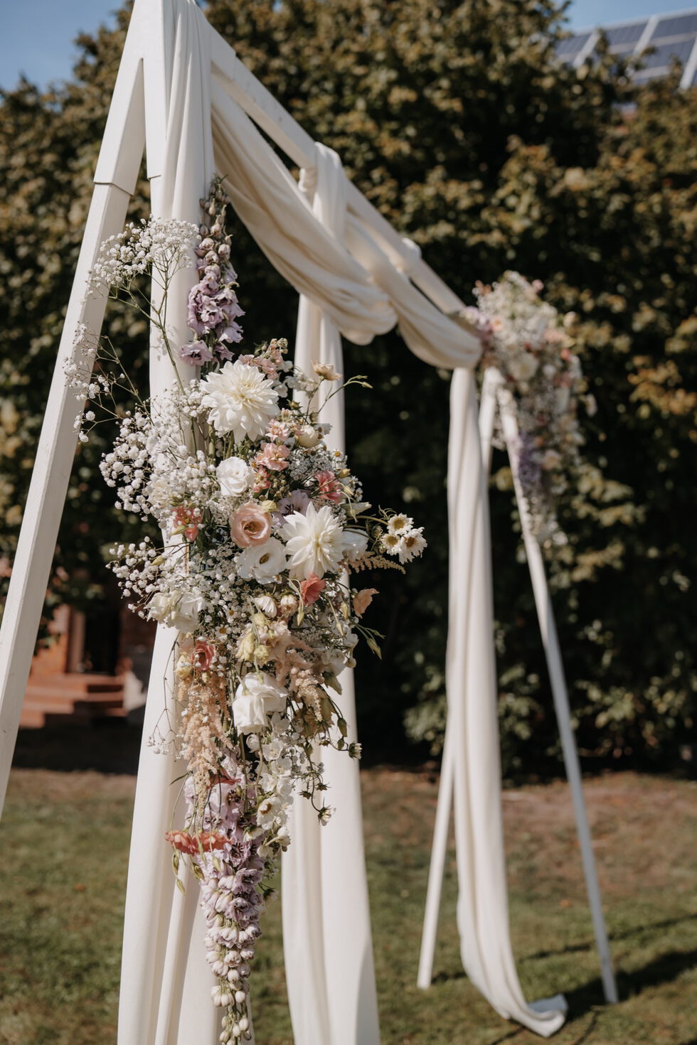 Mit weißen Stoffbahnen und pastellfarbenen Blumen geschmückter Traubogen im Garten.