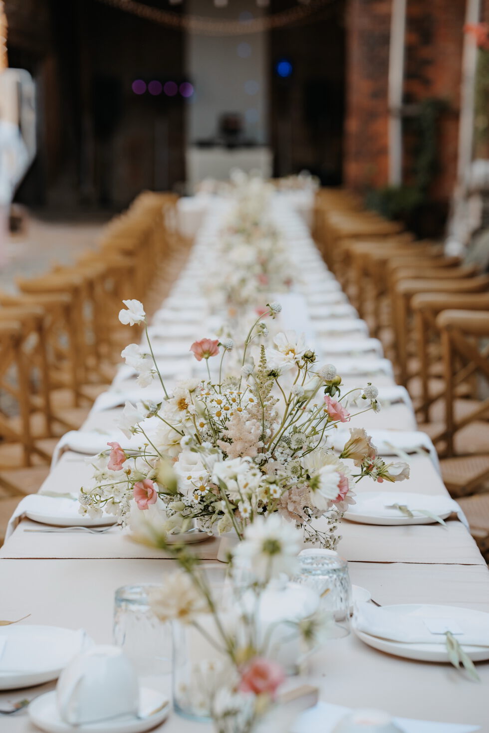 Blumengestecke in Weiß, Creme und zartem Rosé auf einer festlich gedeckten langen Hochzeitstafel in der Scheune.