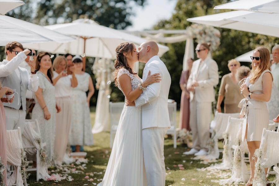 Braut und Bräutigam küssen sich nach der Trauung zwischen ihren Gästen unter weißen Sonnenschirmen.