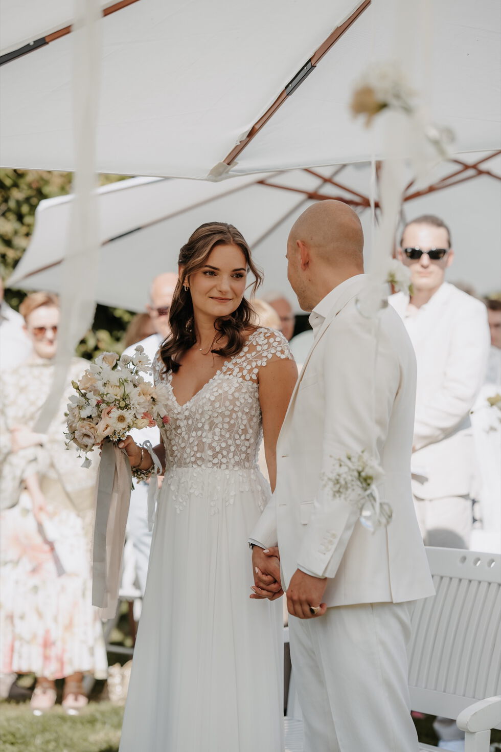 Braut und Bräutigam stehen unter weißen Sonnenschirmen und halten sich während der Trauung an den Händen.
