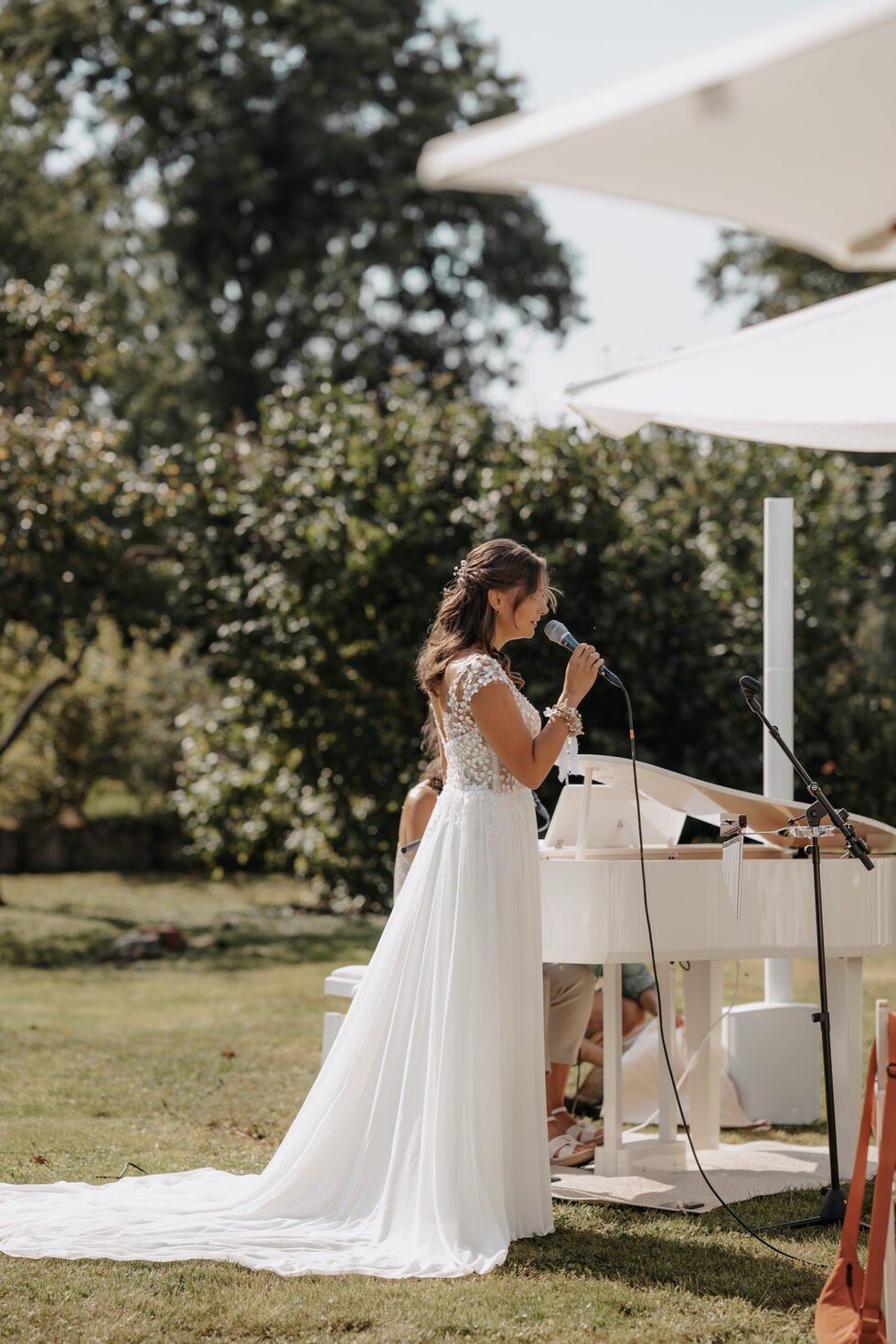 Die Braut singt während der Trauung am Mikrofon, begleitet von einer Pianistin am weißen Flügel im Garten.